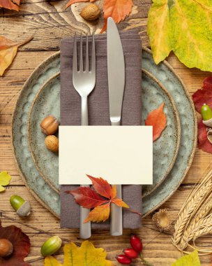 Autumn rustic table place with blank card between colorful leaves and berries, top view, mockup. Flat lay with horizontal card on wooden table,  copy space. Thanksgiving or harvest concept