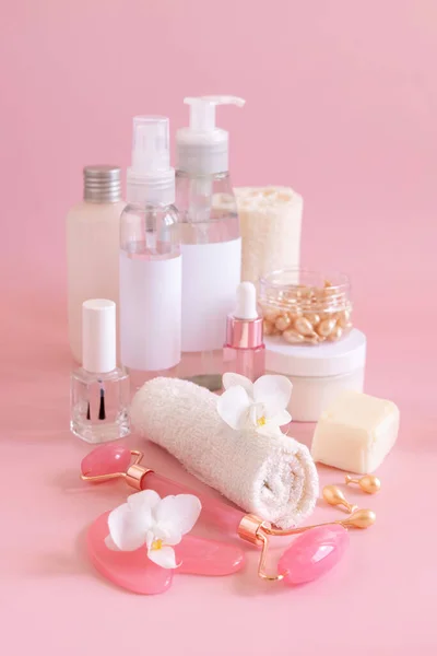 Natural cosmetic jars, skin care accesories and rose quartz face massagers with white orchid flower on light pink close up. Beauty products mockup. Everyday woman face care routine, Spa Treatments