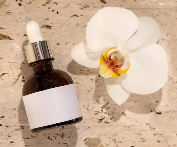 Brown Glass Dropper bottle on stone near white orchid flower top view, label mockup. Skincare handmade beauty product, serum or lotion. Exotic natural cosmetic