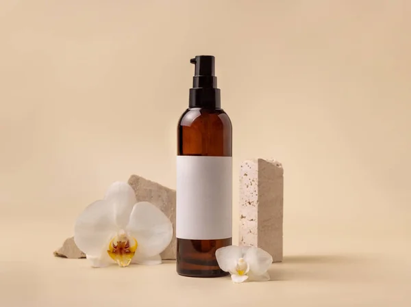 Dark glass cosmetic bottle with blank label near white orchid flowers and stones on light yellow, close up, mockup. Skincare handmade beauty product, tonic or lotion. Exotic natural cosmetic