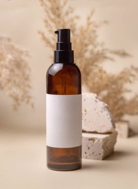 Brown glass cosmetic bottle with blank label near stones and pampas grass on light yellow, close up, mockup. Skincare beauty product. Natural eco friendly cosmetics