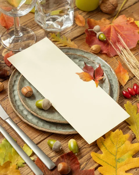 Autumn table setting with red, yellow and orange leaves, berries and blank card close up. Fall Mockup of vertical menu card on wooden table, copy space. Harvest, Halloween and Thanksgiving season