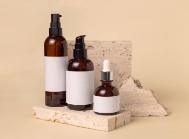 Brown glass one pump bottles on travertine stones on light beige close up, label mockup. Skincare beauty product package. Natural cosmetics, minimal compositio