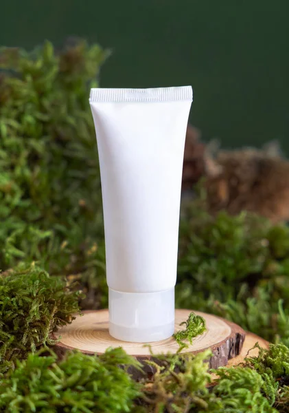 White plastic cream tube on wooden piece between green moss, pine cones and nuts, close up, mockup. Skincare beauty product package. Natural Organic Cosmetic concept.