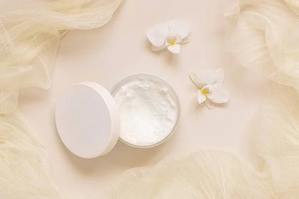 Opened Cream jar with a lid near white orchid flowers and tulle on light yellow top view. Cosmetic Mockup. Exotic natural cosmetics, romantic composition, everyday skincare routin