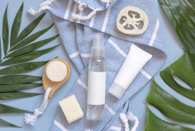 Transparent Refillable spray dispenser bottle and white cream tube on blue bath towel near body care accessories top view, copy space. Packaging mockup. Natural eco friendly Cosmetics