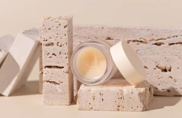 Smass Glass Cosmetic jar on travertine stones on light beige close up. Skincare beauty product package. Exotic natural cosmetics, pastel minimal compositio