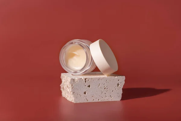 Smass Glass Cosmetic jar on travertine stones on red close up. Skincare beauty product package. Exotic natural cosmetics, pastel minimal compositio