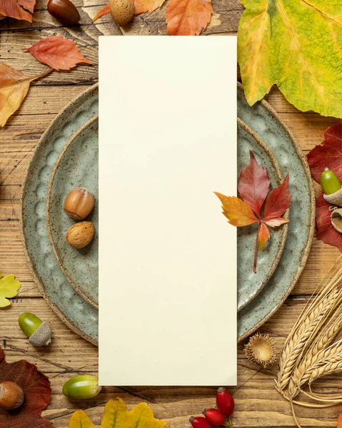 Autumn table setting with red, yellow and orange leaves, berries and blank card top view. Fall Mockup of vertical menu card on wooden table, copy space. Harvest, Halloween and Thanksgiving season