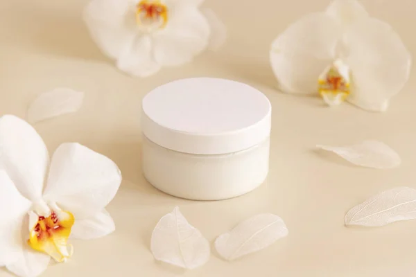 Cream jar with a white lid near white orchid flowers on light yellow close up. Cosmetic packaging Mockup. Exotic natural cosmetics, romantic composition, everyday skincare routin