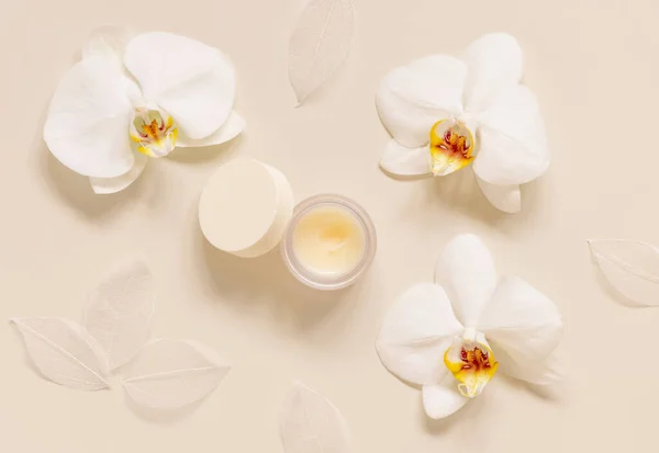 Opened Cream jar with a blank white lid near white orchid flowers on light beige top view. Cosmetic packaging Mockup. Exotic natural cosmetics, romantic composition, everyday skincare routin