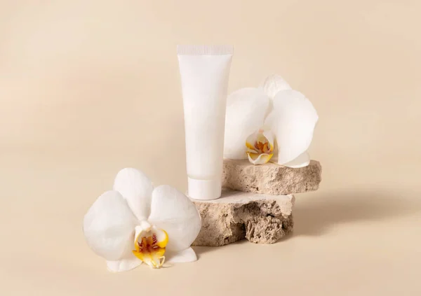 White cream tube on travertine stone near white orchid flower on light yellow, close up, mockup. Skincare handmade beauty product, cream or lotion. Exotic natural cosmetic