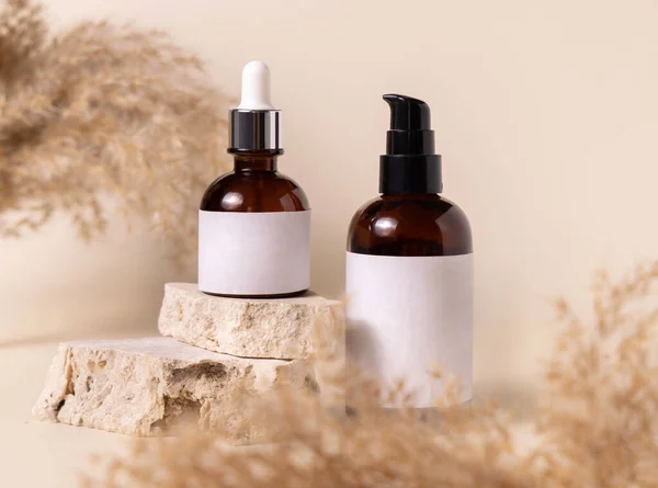 Cosmetic bottles on beige stones near dry pampas grass on light beige close up. Labels Mockup. Skincare beauty product packages. Natural cosmetics, minimal boho compositio