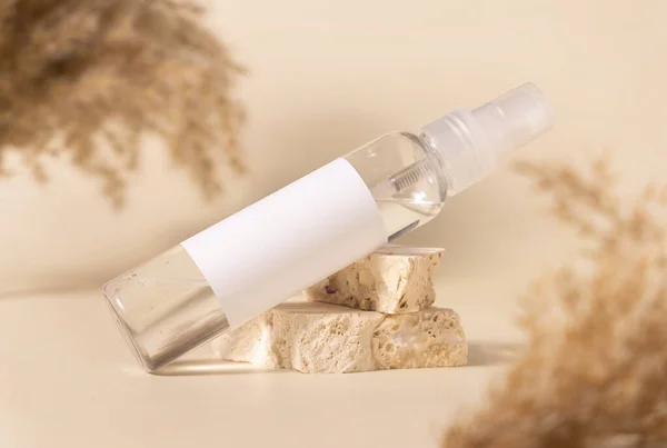 Transparent glass cosmetic bottle with blank label on stones near pampas grass on light yellow, close up, mockup. Skincare beauty product. Natural eco friendly cosmetics