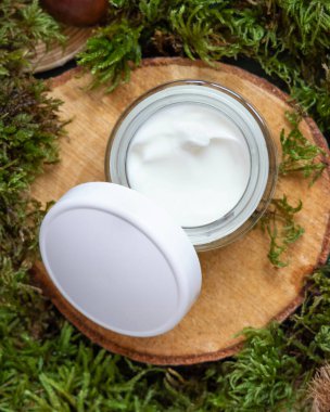 Opened cream jar with white cap on wooden piece between green moss, top view, mockup. Skincare beauty product package. Natural Organic Cosmetic concept
