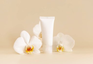 White plastic cream tube near white orchid flowers on light beige close up, mockup. Skincare beauty product, cream or lotion. Exotic natural cosmetics, pastel minimal compositio