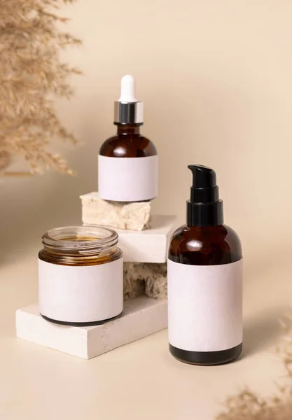 Cosmetic bottles on beige stones near dry pampas grass on light beige close up. Labels Mockup. Skincare beauty product packages. Natural cosmetics, minimal boho compositio