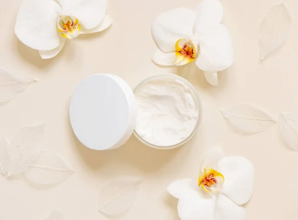 Opened Cream jar with a blank lid near white orchid flowers on light beige top view. Cosmetic Mockup. Exotic natural cosmetics, monochrome minimal flat lay, everyday skincare routin