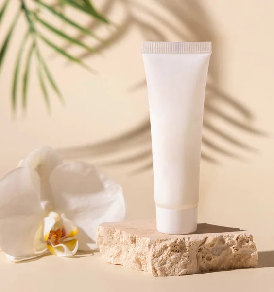 White plastic cream tube on travertine stone near white orchid flower on light beige, Palm leaf hard shadows, close up, mockup. Skincare beauty product. Natural cosmetics. Tropical compositio