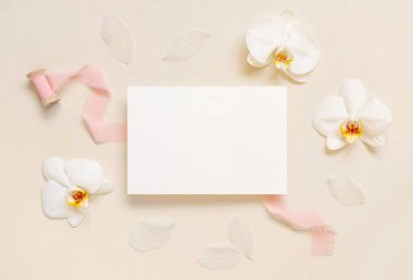 Paper card near white orchid flowers and light pink silk ribbons on light beige, top view, Wedding mockup. Romantic scene with horizontal blank card flat lay. Tropical exotic concept
