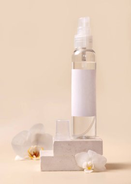Spray dispencer bottle near white orchid flowers and beige stones on light beige close up, mockup. Skincare beauty product package. Natural cosmetics, minimal compositio