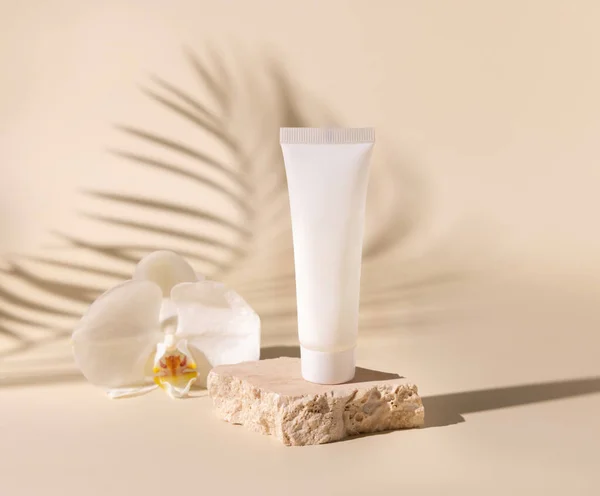 White plastic cream tube on travertine stone near white orchid flower on light beige, Palm leaf hard shadows, close up, mockup. Skincare beauty product. Natural cosmetics. Tropical compositio