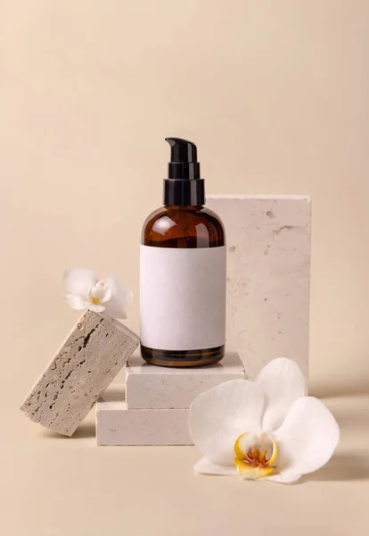 Dark glass one pump cosmetic bottle with blank label near white orchid flowers and stones on light yellow, close up, mockup. Skincare handmade beauty product, tonic or lotion. Exotic natural cosmetic