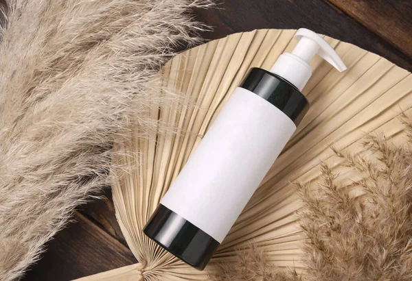Cosmetic one pump bottle on dried palm leaf near pampas grass top view, label mockup. Skincare beauty product, lotion or essence. Bohemian eco friendly cosmetics flat lay with dried leave