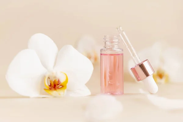 Light Pink opened Dropper Bottle with serum and a pipette cap near white orchid flowers on light beige, close up, copy space. Skincare cosmetic product, essence or serum. Everyday beauty routin