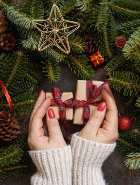 Female hands holding gift boxes near Christmas decorations and fir tree branches top view on a dark table. Christmas holiday atmosphere flat lay. Wrapping presents for loved ones during winter holidays