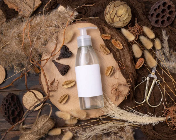 Transparent one pump bottle with liquid on wood near natural boho decorations top view, label mockup. Skincare beauty product, lotion or essence. Bohemian eco friendly cosmetics flat lay with dried leave
