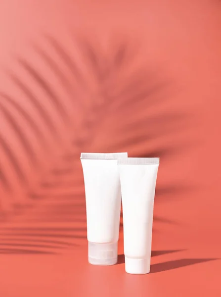 Two white blank plastic tubes on pink close up, palm leaf hard shadows, mockup. Skincare beauty product, body lotion or cream. Natural cosmetic