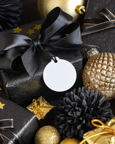 Black and golden Christmas Gift Boxes with a bow and ornaments close up, round paper gift tag mockup, copy space. Dark winter composition with blank label card for Christmas, New Year, Birthday, Anniversary