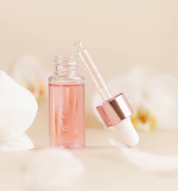 Light Pink opened Dropper Bottle with serum and a pipette cap near white orchid flowers on light beige, close up, copy space. Skincare cosmetic product, essence or serum. Everyday beauty routin
