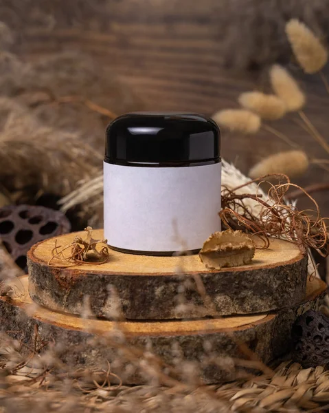 Cream jar with blank label on wood near natural decorations close up, package mockup. Eco friendly skincare product, lotion or cream. Bohemian composition with dried leaves and flower