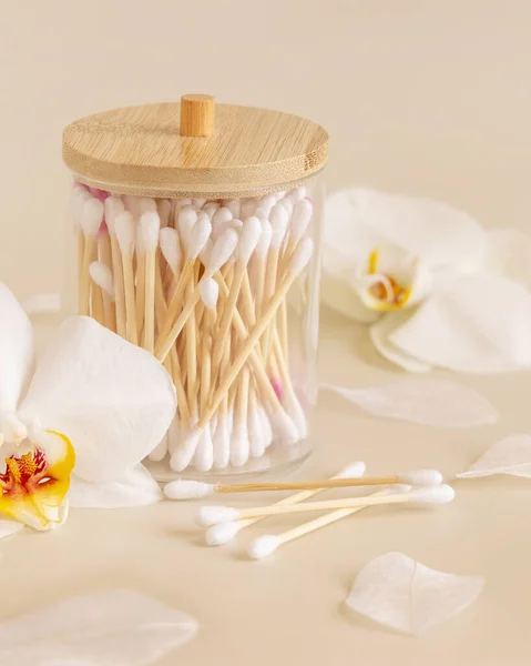 Bamboo cotton buds in a jar near white orchid flowers on light beige, close up. Organic skincare product for everyday beauty routine. Eco friendly Biodegradable cotton swab
