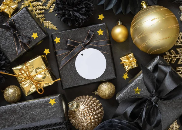 Black and golden Christmas Gift Boxes with a bow and ornaments close up, round paper gift tag mockup, copy space. Dark winter composition with blank label card for Christmas, New Year, Birthday, Anniversary
