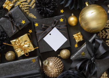 Üzerinde fiyonk ve süs bulunan siyah ve altın Noel hediye kutuları. Kare kağıt hediye etiketi maketi, kopyalama alanı. Noel, Yeni Yıl, Doğum Günü, Yıldönümü için boş etiket kartıyla karanlık kış kompozisyonu 
