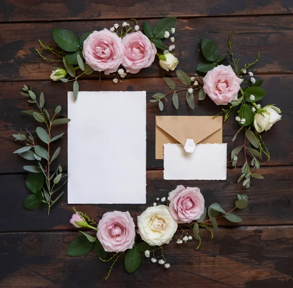 Cards and envelope between pink and cream roses on brown wood top view,  wedding mockup. Romantic scene with cards and pastel flowers flat lay. Valentines, Spring or Mothers day concept