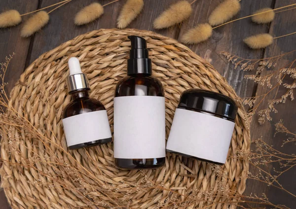 Cosmetic bottles on wattled boho placemat on wood near Hare's Tail grass top view, packaging label mockup. Natural skincare product for everyday beauty routine. Bohemian and eco friendly