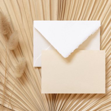 Tavşan 'ın kuyruğundaki çimlerin yanındaki kuru bej palmiye yaprağının üzerindeki boş kart ve zarf, maket. Yatay kartlı Boho sahnesi. Bohem ya da doğaya yakın tebrik kartı ya da düğün davetiyesi