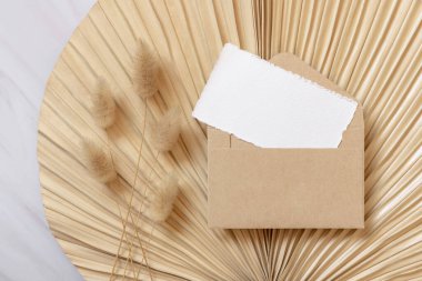 Tavşan 'ın kuyruğundaki çimlerin yanındaki kuru bej palmiye yaprağının üzerindeki boş kart ve zarf, maket. Yatay kartlı Boho sahnesi. Bohem ya da doğaya yakın tebrik kartı ya da düğün davetiyesi