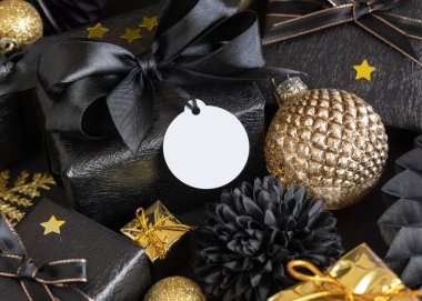 Black and golden Christmas Gift Boxes with a bow and ornaments close up, round paper gift tag mockup, copy space. Dark winter composition with blank label card for Christmas, New Year, Birthday, Anniversary 