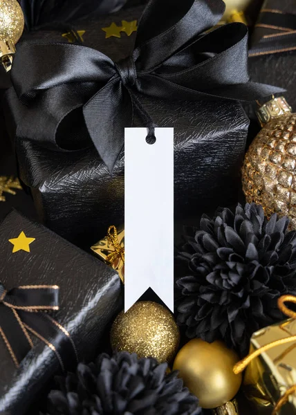 Black and golden Christmas Gift Boxes with a bow and ornaments close up, vertical paper gift tag mockup, copy space. Dark winter composition with blank label card for Christmas, New Year, Birthday, Anniversary