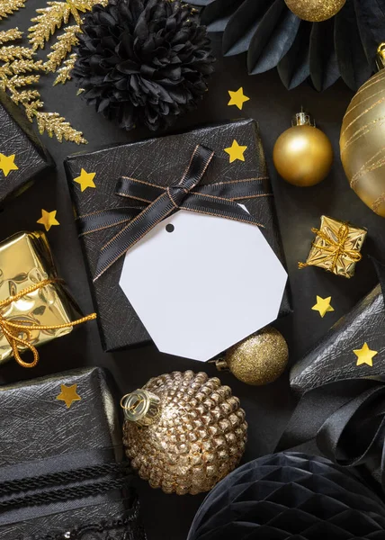 Black and golden Christmas Gift Boxes with a bow and ornaments close up, square paper gift tag mockup, copy space. Dark winter composition with blank label card for Christmas, New Year, Birthday, Anniversary