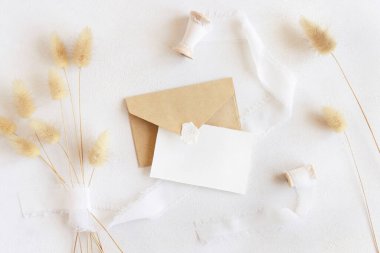 İpek kurdelelerin yanındaki boş kart ve zarf ve kurutulmuş tavşanın kuyruk çimlerinin beyaz, sahte görünümü. Yatay kartlı Boho romantik sahnesi. Bohem ya da doğaya yakın selamlama ya da düğün davetiyesi