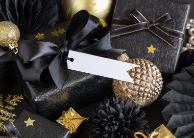 Black and golden Christmas Gift Boxes with a bow and ornaments close up, horizontal paper gift tag mockup, copy space. Dark winter composition with blank label card for Christmas, New Year, Birthday, Anniversary 
