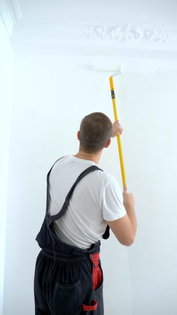 Young man paints the wall in the room with a roller white paint, vertical — Stock Video
