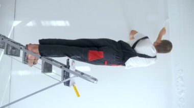 rear view of a man in overalls painting a wall with white paint while standing on a stepladder. vertical video