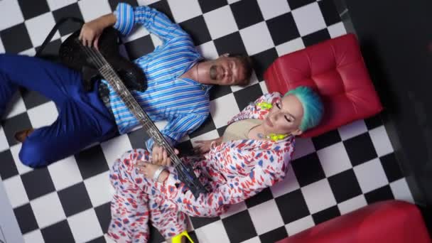 A couple, a man playing the electric guitar and a travesty actor lie on the floor with a checkerboard pattern. rotating image — Stockvideo